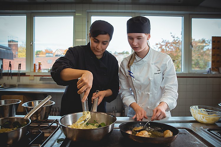 Bild på två elever som lagar mat vid en spis i köket på Restaurang- och livsmedelsprogrammet. 